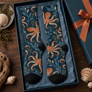 Puede incluir: Un par de calcetines con fondo azul oscuro, un patrón de pulpos naranjas y algas azul claro. Los calcetines tienen las punteras y los talones de color negro sólido y se presentan dentro de una caja de regalo azul oscuro con papel de seda.