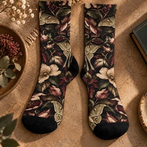 May include: A pair of crew socks featuring a dark black background with a botanical print of pale green moths, white flowers, deep red berries, and dark green foliage. The socks have solid black toe and heel sections with a seamless knit construction.