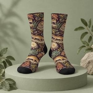 May include: A pair of crew-length socks featuring a dark purple background with a repeating pattern of yellow and brown leopard geckos, white lotus flowers, green succulents, and various rocks. The socks have solid black toe and heel sections.