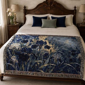 May include: A rectangular woven throw blanket featuring a dark blue and gold floral pattern of irises and foliage. The blanket has a decorative fringe along all four edges and is draped across the foot of a bed with neutral bedding.
