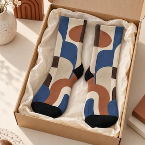 May include: A pair of crew socks featuring a geometric pattern of interlocking arches and circles in shades of blue, terracotta, cream, and black. The socks are presented inside a cardboard gift box lined with white tissue paper.