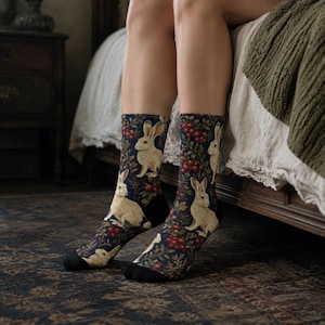May include: A pair of crew socks featuring a dark navy blue background with a repeating pattern of cream-colored rabbits, red berries, and green foliage. The socks have solid black toe and heel sections, providing a distinct contrast to the print.