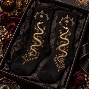 Könnte beinhalten: Ein Paar schwarze Socken mit einer detaillierten goldfarbenen Schlangengrafik, die sich um ein vertikales Element windet, gekrönt von einem verzierten kreisförmigen Rahmen und einem Sonnensymbol. Das Design enthält filigrane Muster in Gold und Dunkelrot.