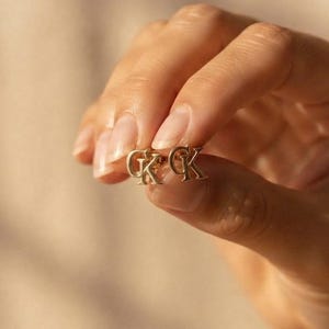 May include: A pair of gold-toned stud earrings featuring the letters CK in a serif font. The earrings have a polished metallic finish and are designed as small, compact initials. Each earring displays the letters C and K joined together.
