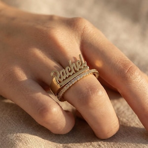 May include: A gold-toned ring featuring a nameplate with the name Rachel in a cursive script font, positioned directly above a second, thinner band that is encrusted with a row of small, clear, sparkling stones along its entire outer surface.