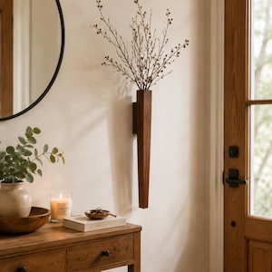 Peut inclure: Un vase mural en bois brun foncé, haut et étroit, au profil rectangulaire. Le vase est fixé verticalement sur un mur de couleur claire et contient un bouquet de fines brindilles nues et ramifiées avec de petits bourgeons aux extrémités.