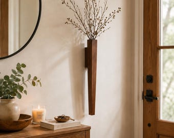 May include: A tall, narrow, dark brown wooden wall-mounted vase with a rectangular profile. The vase is mounted vertically on a light-colored wall and holds a cluster of thin, bare, branching twigs with small buds at the tips of the stems.