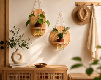 May include: Two wall-mounted wooden plant holders with glass vases, one oval and one circular, each suspended by a white rope. A beige ceramic donut-shaped vase with green branches sits on a wooden cabinet with cane doors below the wall decor.