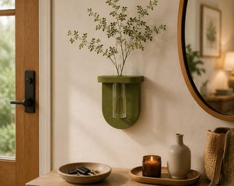 May include: A wall-mounted olive green semi-circular shelf featuring a central cylindrical glass tube holding a leafy green plant stem. The shelf is constructed with a smooth, matte finish and provides a vertical display for botanical elements.