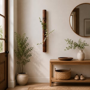 May include: A dark brown cylindrical bamboo wall vase featuring three circular openings along its length, holding a delicate green leafy branch with small white flowers. The bamboo piece is mounted vertically on a plain light-colored wall.