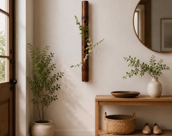 May include: A dark brown cylindrical bamboo wall vase featuring three circular openings along its length, holding a delicate green leafy branch with small white flowers. The bamboo piece is mounted vertically on a plain light-colored wall.