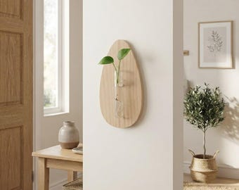 May include: A wall-mounted light wood plaque shaped like a teardrop featuring a clear glass test tube vase. A small green plant cutting with two leaves is placed inside the glass tube, which is secured to the center of the wooden surface.