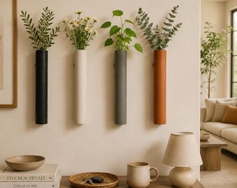 May include: Four cylindrical wall-mounted vases in black, off-white, gray, and terracotta, each holding green foliage or white flowers. The vases are arranged in a horizontal row on a plain wall above a wooden console table with decor items.