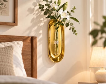 May include: A gold-colored oval wall-mounted metal plaque featuring a central clear glass test tube vase. The vase holds a bouquet of white lily flowers and green eucalyptus stems, creating a vertical floral display against the wall surface.