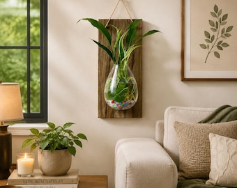 May include: A wall-mounted planter featuring a rectangular dark wood board with a teardrop-shaped glass vase attached. The vase contains a green plant and colorful pebbles at the bottom. A thin rope is attached to the top for hanging on the wall.