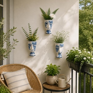 Può includere: Tre vasi da parete in ceramica bianca, ciascuno a forma di volto umano con tratti astratti dipinti in blu. I vasi sono disposti verticalmente e diagonalmente su una parete, contenendo ciascuno fogliame verde e piccoli fiori bianchi.