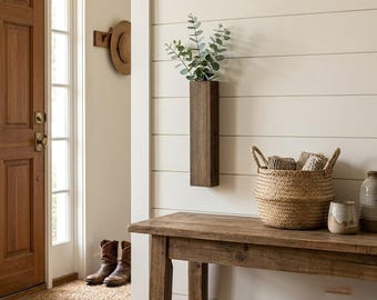 May include: A rectangular dark wood wall-mounted vase holding green eucalyptus stems, positioned above a wooden console table with a woven seagrass basket, a ceramic jar, and a glass vessel, set against white horizontal shiplap wall paneling.