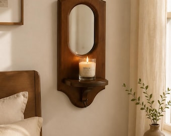 May include: A wall-mounted wooden mirror featuring an arched top and a small integrated shelf. A glass candle holder with a lit white candle sits on the shelf. The mirror frame has a smooth, dark brown finish and a curved, decorative silhouette.