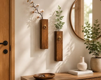 May include: Two rectangular dark brown wooden wall-mounted vases, each featuring a small gold ring accent. One vase holds a branch with white blossoms, while the other contains a green leafy stem. The vases are mounted on a plain wall.