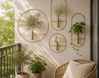 May include: Four gold-toned metal wall-mounted planters with cylindrical vases. Two are circular and two are oval, each holding various green plants and white flowers. The planters are arranged on a wall above a small wooden table and a wicker chair.