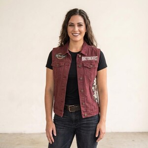 Può includere: Un gilet in denim bordeaux con finitura consumata, chiusura frontale con bottoni e colletto. Il gilet presenta una toppa circolare sul petto destro, la scritta BIKETOBERFEST sul petto sinistro e un motivo floreale ricamato sul lato sinistro.