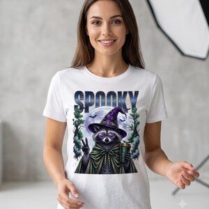 May include: A white short-sleeved t-shirt featuring a graphic of a raccoon wearing a purple witch hat, heart-shaped glasses, and a green cape. The raccoon holds a coffee cup and makes a peace sign. The text SPOOKY appears above the raccoon.