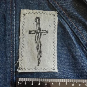 Op de afbeelding: Een rechthoekige lichtgrijze stoffen patch met een zwarte inktillustratie van een kruis, vastgenaaid op een donkerblauw spijkerjack. Een zwarte plastic liniaal is onder de patch geplaatst en toont een breedte van 5 centimeter.