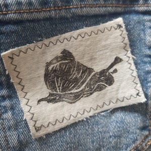 Könnte beinhalten: Ein rechteckiger, cremefarbener Stoffaufnäher mit einer schwarzen, mit Tinte gedruckten Schneckenillustration, der mit einem dunklen Faden und einem Zickzackstichmuster entlang aller vier Kanten auf einer blauen Jeanstasche befestigt ist.