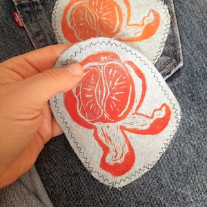 May include: Two circular light blue denim fabric patches featuring a hand-printed orange citrus fruit design in vibrant red-orange ink. Each patch has a dark grey zigzag stitched border and is displayed against a dark blue denim fabric background.