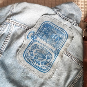 May include: A light blue denim jacket featuring a rectangular fabric patch stitched onto the back. The patch displays a blue ink print of an open sardine tin with the text SARDINES IN OLIVE OIL and a central fish illustration.