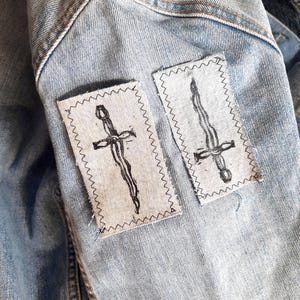 May include: Two rectangular light-wash denim patches are sewn onto a light blue denim jacket. Each patch features a black ink illustration of a wavy-bladed dagger or sword. The patches are attached with a visible black zigzag stitch border.