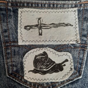 May include: A pair of blue denim jeans featuring two rectangular fabric patches sewn onto the back pocket. The top patch displays a black ink illustration of a sword, while the bottom patch features a black ink illustration of a snail.
