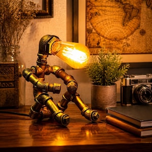 May include: A table lamp constructed from dark metallic industrial pipes, shaped like a seated figure. The lamp features a glowing, warm-toned Edison-style light bulb as the head, with visible threaded joints and a weathered, textured finish.