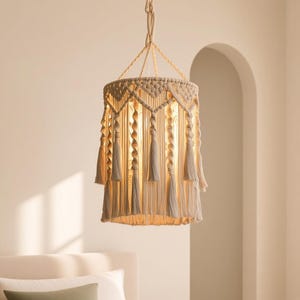 May include: A beige macrame pendant light fixture featuring a cylindrical shade with intricate knotted patterns, twisted cord accents, and hanging fringe tassels. The lamp is suspended by a braided cord and emits a warm, soft glow from within.
