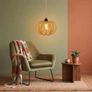 May include: A pumpkin-shaped woven pendant light with a warm glow hangs above an olive green velvet armchair draped with a patterned throw blanket. A small potted green plant and a ceramic mug sit on a round wooden side table nearby.