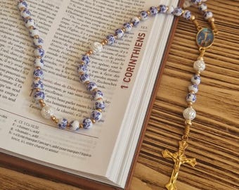 Può includere: Un rosario con perline a motivo floreale blu e bianco alternate a perline bianche effetto perla, collegate da maglie in metallo dorato. Include un elemento centrale circolare blu con un'immagine religiosa e un crocifisso color oro.