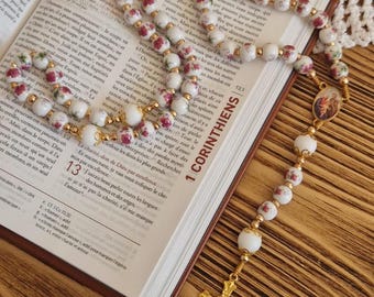 May include: A rosary featuring white beads decorated with small pink floral patterns, connected by gold-toned metal links. The rosary includes a central oval medal depicting a religious figure and a gold-toned crucifix at the bottom end.