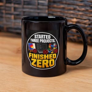 May include: A black ceramic mug featuring a circular graphic with the text STARTED THREE PROJECTS, FINISHED ZERO. The design includes illustrations of a painting on an easel, a laptop, and a building block structure, each marked with a red X.