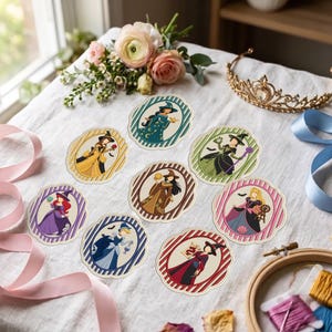 Könnte beinhalten: Ein Set aus acht runden Aufklebern mit Illustrationen von als Hexen verkleideten Figuren, jeweils in einem gewellten Rahmen mit verschiedenen Farbschemata und Mustern, angeordnet auf einem weißen Stoff mit einem goldenen Diadem und rosa Band.