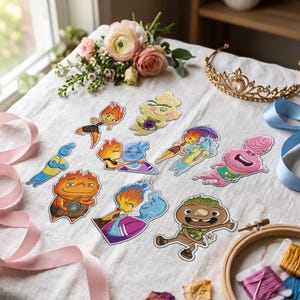 Peut inclure: Une collection d'écussons brodés représentant des personnages de dessins animés colorés, incluant des figures de flammes, un personnage bleu et une créature brune, disposés sur un tissu blanc avec un diadème doré et un ruban rose.
