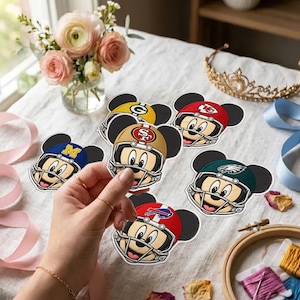 Op de afbeelding: Een verzameling gestanste stickers met Mickey Mouse in verschillende football-teamhelmen, waaronder de 49ers, Packers, Chiefs, Eagles, Bills en een team met een geel M-logo, weergegeven op een witte ondergrond met bloemaccenten.