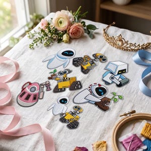 May include: A collection of embroidered patches featuring characters from the movie WALL-E, including WALL-E and EVE in various poses, a pink robot, and a white robot, arranged on a white fabric surface with ribbons and a gold tiara.