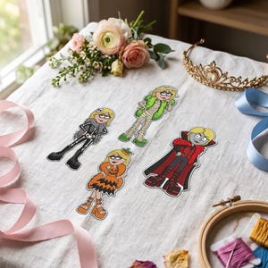 May include: Four cartoon-style stickers featuring characters in Halloween costumes: a skeleton, a mummy with green accents, a pumpkin-themed dress, and a vampire with a red cape. Each character has blonde hair and is depicted in a flat, graphic design.