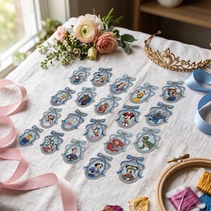 Peut inclure: Une collection de vingt et un écussons brodés représentant divers personnages de dessins animés, chacun encadré par une bordure bleue décorative avec un nœud au sommet. Les écussons sont disposés sur un tissu blanc avec des rubans roses et un diadème.