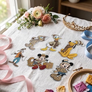 May include: A collection of six embroidered iron-on patches featuring cartoon characters dressed in winter clothing, including Mickey Mouse, Minnie Mouse, Donald Duck, Goofy, and Pluto, arranged on a white fabric surface with floral accents.