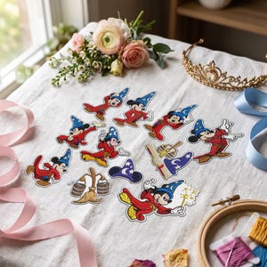 Può includere: Una collezione di toppe ricamate raffiguranti Topolino come stregone con tunica rossa e cappello blu, insieme a una scopa che trasporta secchi, un libro di incantesimi e un cappello da stregone separato, disposti su un tessuto bianco.