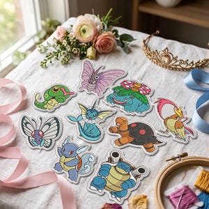 May include: A collection of nine colourful embroidered patches featuring various fictional creatures, including a green caterpillar, a purple butterfly, a blue turtle-like creature, and a bird, arranged on a white fabric surface with ribbons.