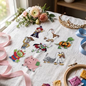 Pode incluir: Uma coleção de patches bordados com vários personagens de desenhos animados em fantasias de Halloween, incluindo um patrulheiro espacial, um cowboy, um porco, um dinossauro verde numa abóbora e um cão com corpo de mola, sobre tecido branco.
