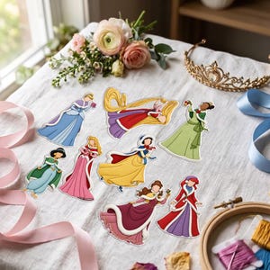 May include: A collection of eight die-cut stickers featuring female characters in winter-themed gowns, including blue, red, yellow, green, and purple dresses with white fur trim. The stickers are arranged on a white fabric surface with ribbons.