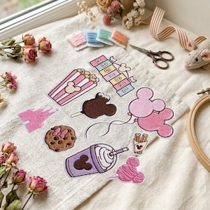 May include: A collection of embroidered iron-on patches featuring a pink castle, popcorn bucket, chocolate-covered treat, cookie, purple drink, churros, cotton candy, balloons, and a banner with the text MAGICAL, all arranged on a beige fabric.